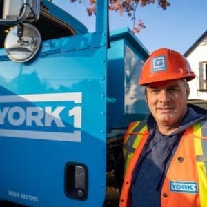 A confident, smiling employee standing next to a blue YORK1 roll-off truck with a dumpster bin.